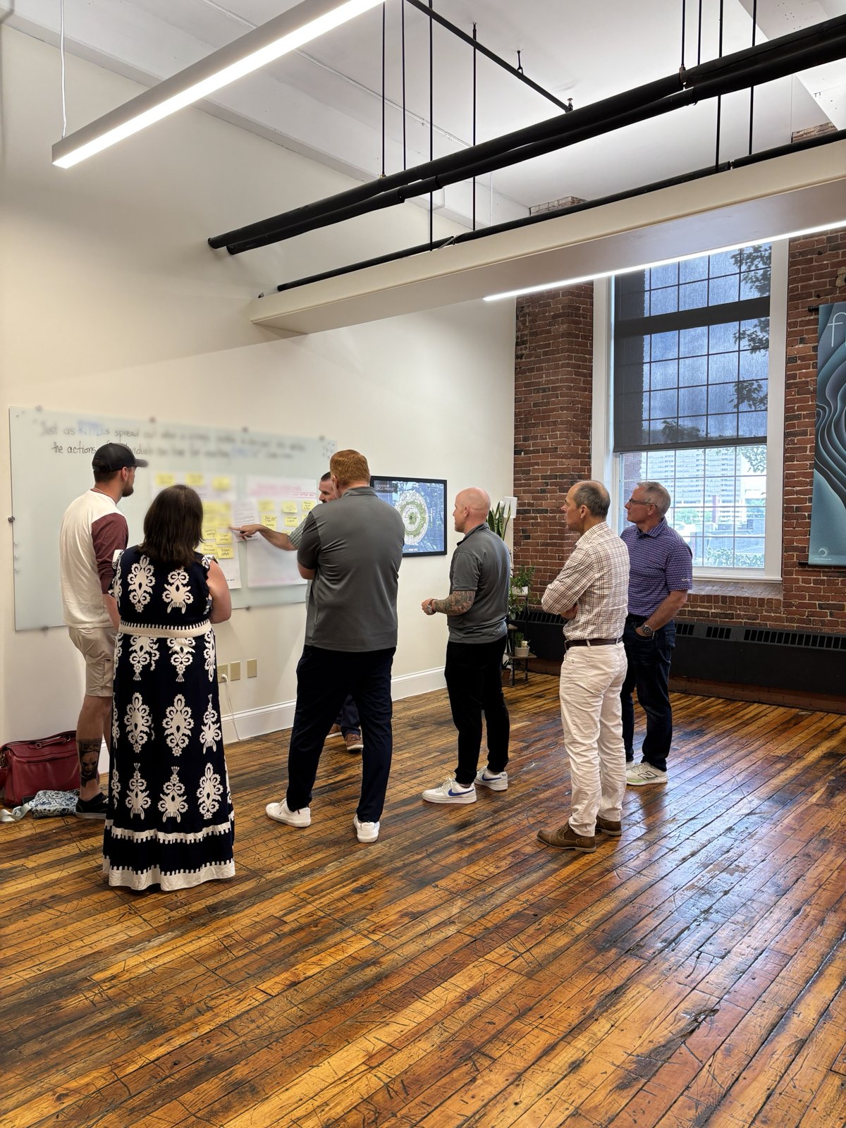 A leadership team on their feet at a wall of sticky notes in a brick mill space, working through ideas together