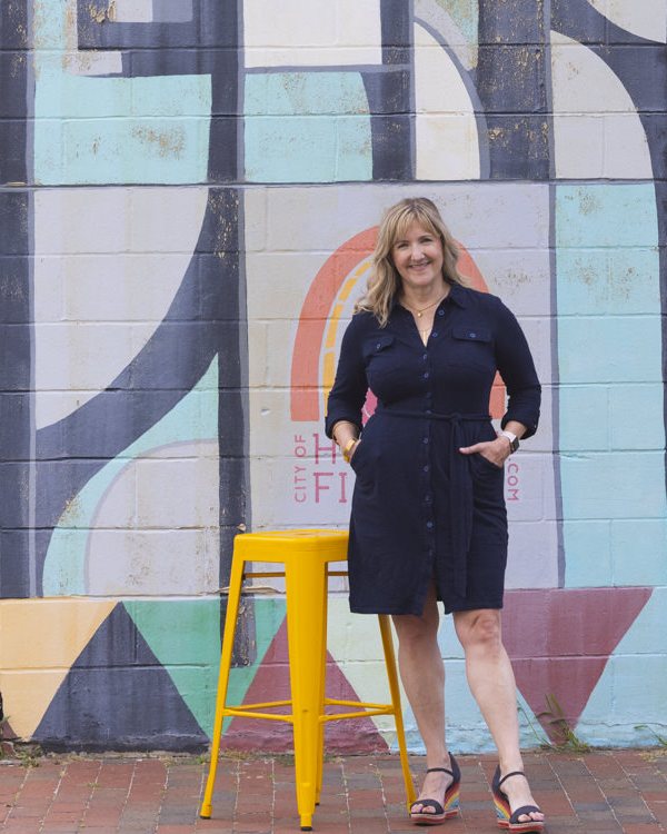 Justine Bassett standing in front of a colorful painted mural beside a yellow stool
