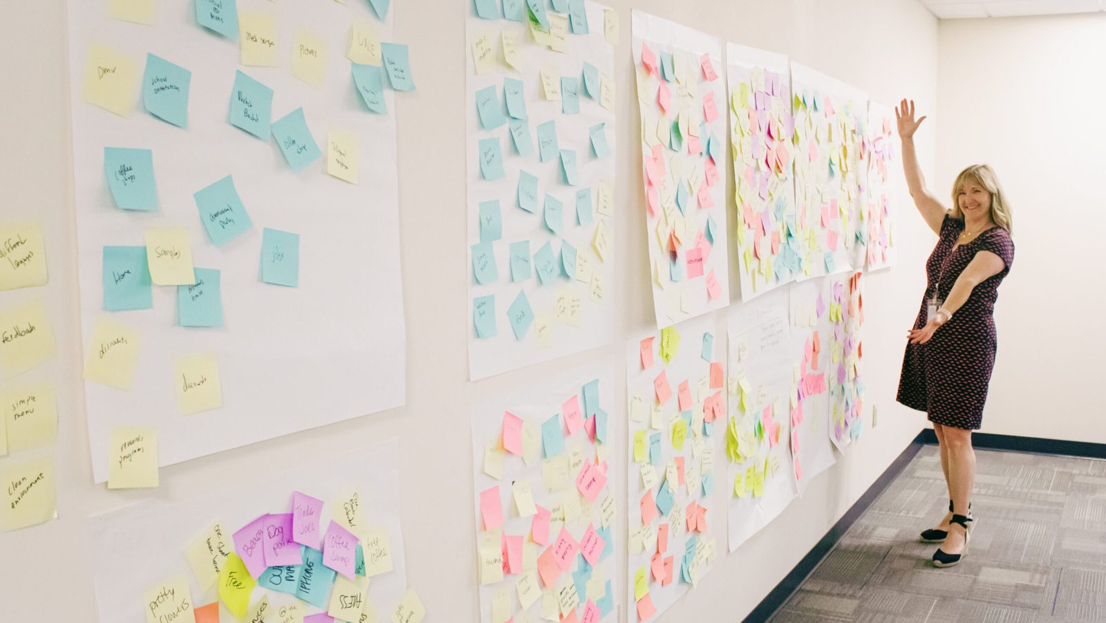 Justine standing beside a long wall covered in colorful sticky notes from a workshop, arms raised in celebration